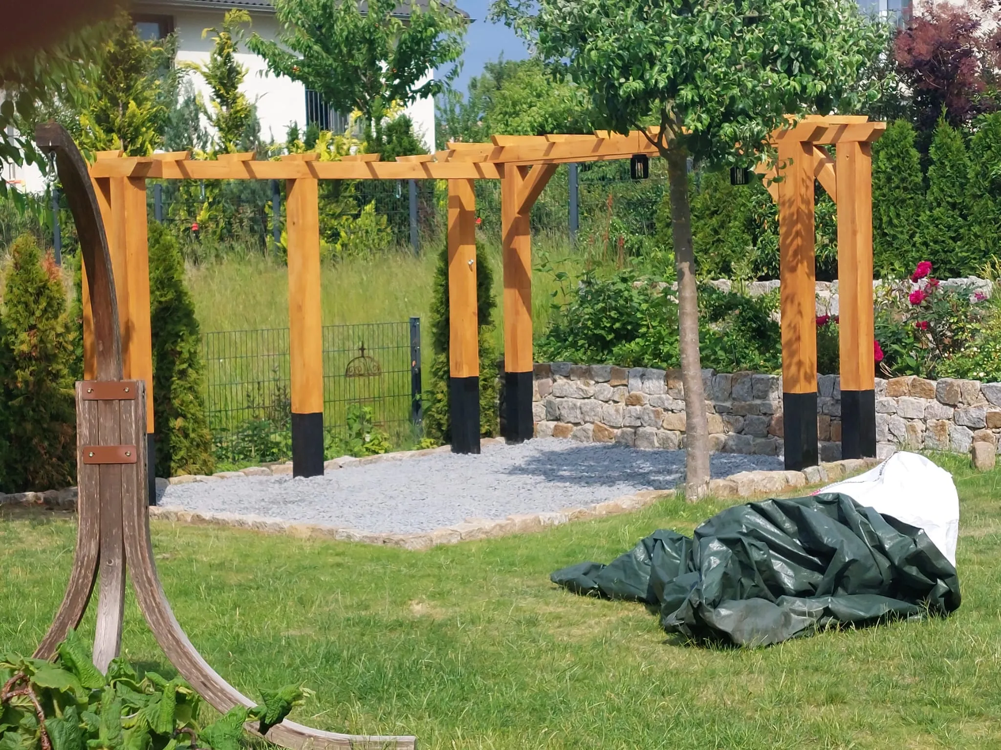 Holzpergola über einer Schotterfläche, dahinter eine Natursteinmauer